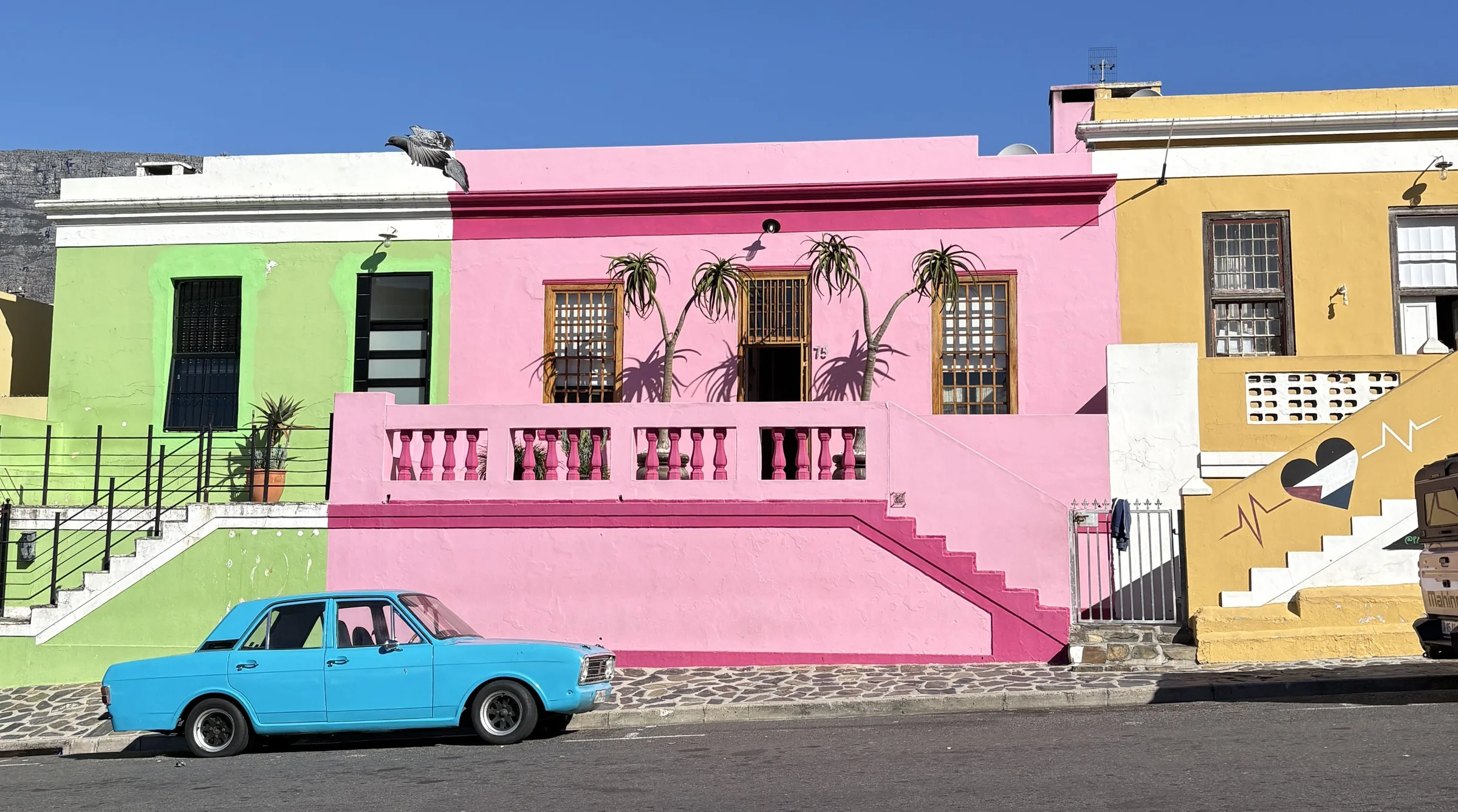 A row of Bo-Kaap houses in pink, green and ochre along a steep Cape Town street, a turquoise vintage car parked at the kerb.