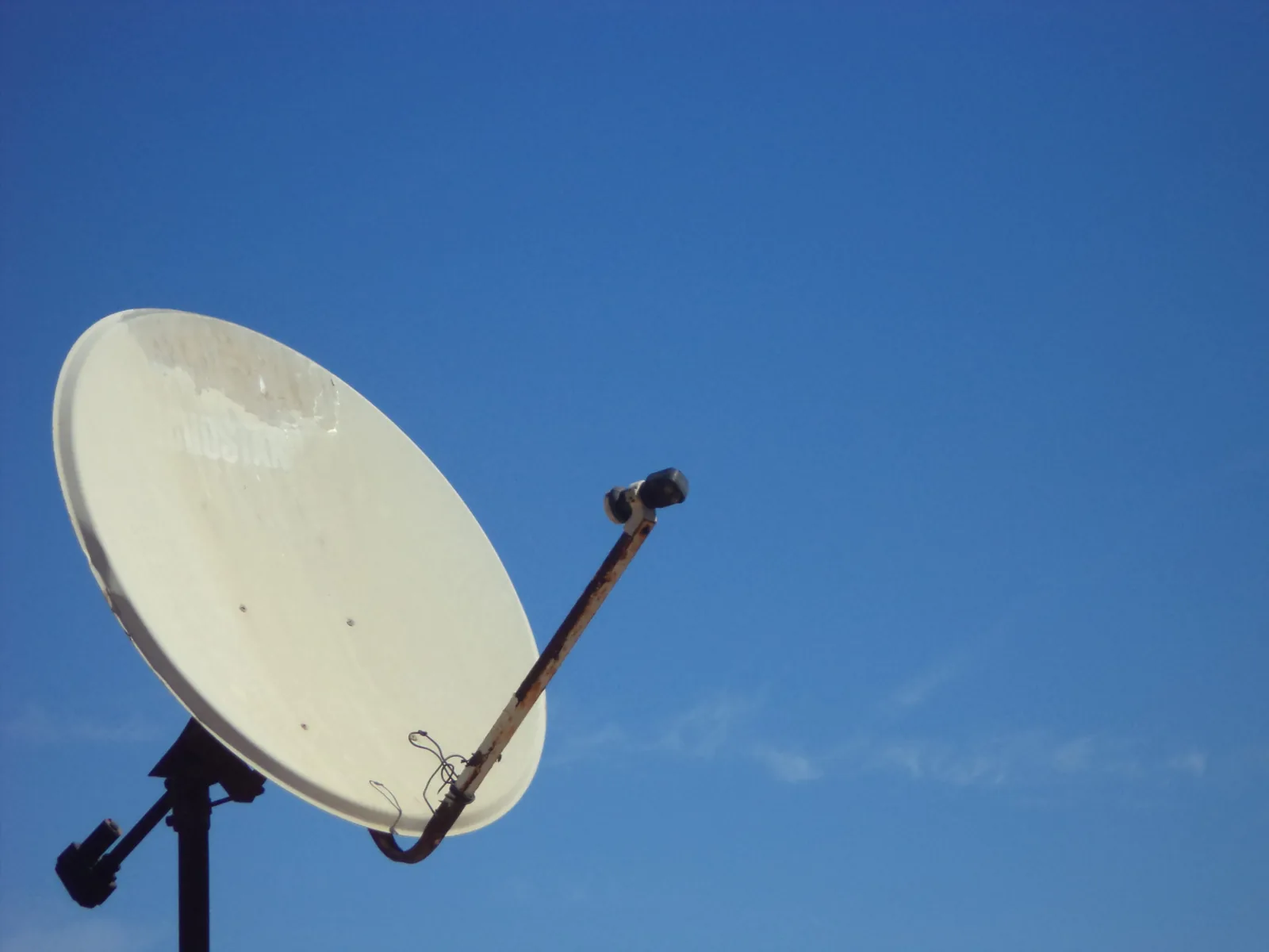 A weathered cream satellite dish on a steel bracket against a clear blue sky, cabling looped back to the LNB arm.