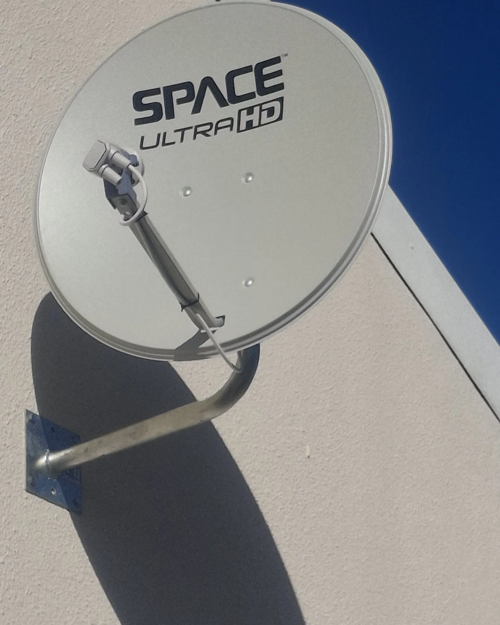 A satellite dish bracketed to a white rendered wall, throwing a hard-edged shadow across it in full afternoon sun.